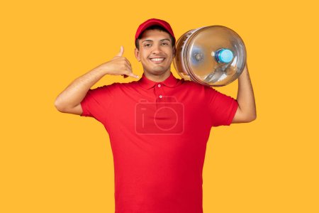 El repartidor con una camisa roja sostiene una enorme botella de agua en su mano y muestra el pulgar hacia arriba en el fondo amarillo del estudio.