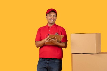 A cheerful delivery guy in a red uniform holds a clipboard while standing beside cardboard boxes. The bright yellow background highlights his friendly demeanor as he prepares for a package delivery.