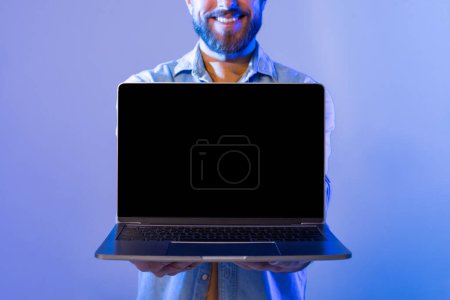 A smiling man with a beard stands against a blue background, holding a laptop with the blank screen facing forward. He appears cheerful and enthusiastic, cropped