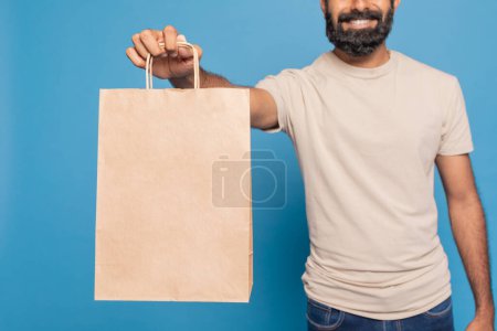 Foto de Recortado de alegre repartidor con barba está sosteniendo una bolsa de papel marrón en blanco delante de un fondo azul brillante - Imagen libre de derechos