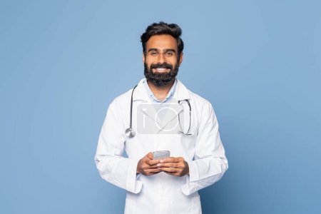 Un médico indio alegre con una bata blanca de laboratorio y estetoscopio se levanta sobre un fondo azul, sonriendo mientras interactúa con su teléfono inteligente