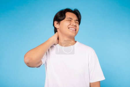 Un jeune Asiatique en t-shirt blanc souffre visiblement de douleurs au cou. Il tient son cou avec sa main droite, exprimant son inconfort. La toile de fond est d'une couleur bleu uni