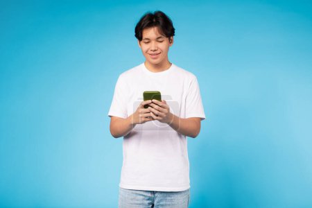 Chico asiático vestido con una camisa blanca está absorto en su teléfono celular, centrándose intensamente en la pantalla. Parece estar desplazándose o leyendo algo en el dispositivo, con una expresión seria