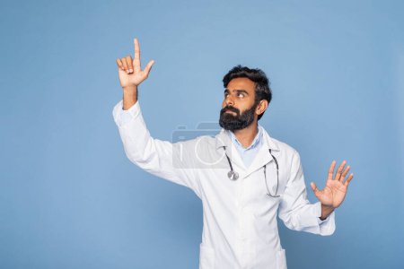 Ein bärtiger indischer Arzt mit weißem Laborkittel und Stethoskop gestikuliert mit der rechten Hand, als würde er auf etwas zeigen, vor einem festen blauen Hintergrund.