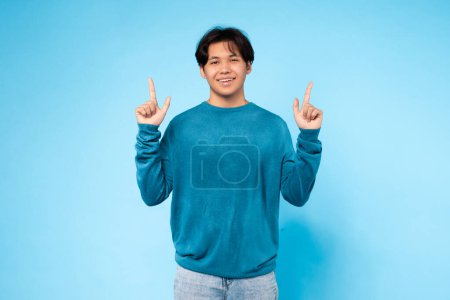 Asiatischer fröhlicher junger Mann in blauem Pullover und Jeans steht vor einem dazu passenden blauen Hintergrund. Er lächelt und zeigt mit beiden Händen nach oben.
