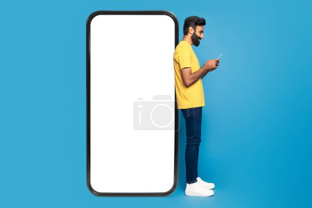 Un joven indio que lleva una camisa amarilla y jeans está absorto en el uso de su teléfono inteligente. Él está parado contra un marco gigante del teléfono con una pantalla blanca en blanco, fijada contra un fondo azul vivo