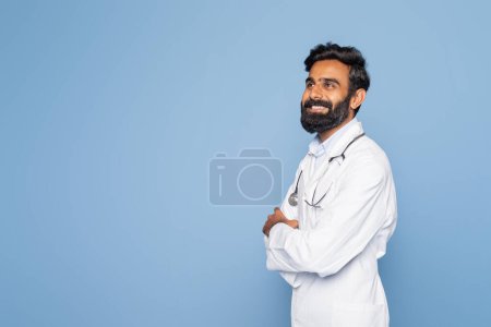 A bearded Indian doctor wearing a white coat and stethoscope smiles while standing confidently against a blue studio background. His arms are crossed, conveying professionalism and warmth.