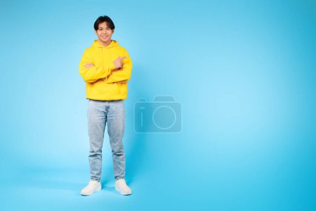 Asian guy wearing a yellow hoodie is standing with his arms crossed, pointing at copy space. He appears confident and assertive.