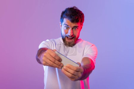A man with a beard and wearing a white shirt is enthusiastically playing a game on his smartphone. The room is lit with vibrant pink and purple lighting, indicating it is nighttime.