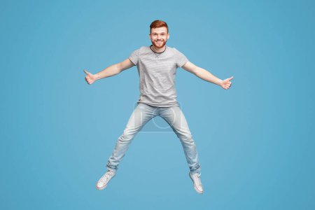 junger rothaariger Millennial-Typ springt in die Luft auf blauem Hintergrund, zeigt Daumen hoch Geste, volle Länge, Studio, leerer Raum