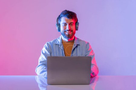 A man wearing headphones is sitting at a desk with a laptop in front of him. He appears to be enjoying music while working, with a smile on his face. The room is illuminated