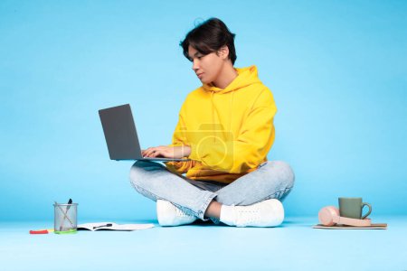 Asiatischer junger Mann mit gelbem Kapuzenpullover und hellblauer Jeans sitzt im Schneidersitz auf dem Boden und konzentriert auf seinen Laptop. Er ist von Lernmaterialien umgeben