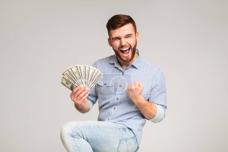 Win a lottery. Young handsome guy happy to have fan of dollars in hand, grey studio background, free space