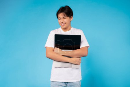 Asian young guy wearing a white t-shirt and jeans is standing against a vibrant blue background. He is smiling and appears content while holding a laptop close to his chest.