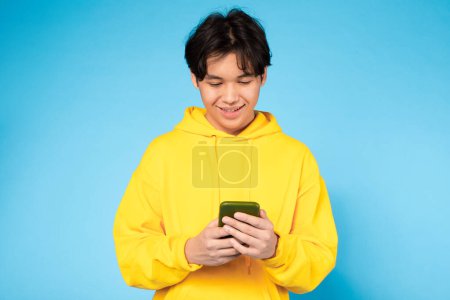 asiático chico usando un amarillo sudadera con capucha se centra en su teléfono celular pantalla, absorto en lo que él está viendo o leyendo.