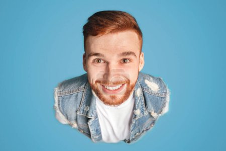 Above portrait of funny millennial redhaired guy, smiling upwards at camera, blue background, studio