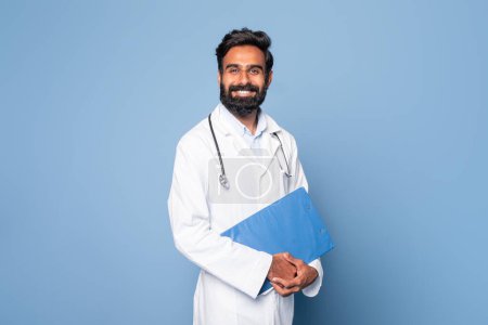 Ein fröhlicher indischer Arzt ist zu sehen, wie er einen blauen Ordner in der Hand hält, während er vor einem schlichten blauen Hintergrund steht. Er trägt einen weißen Laborkittel und ein Stethoskop um den Hals
