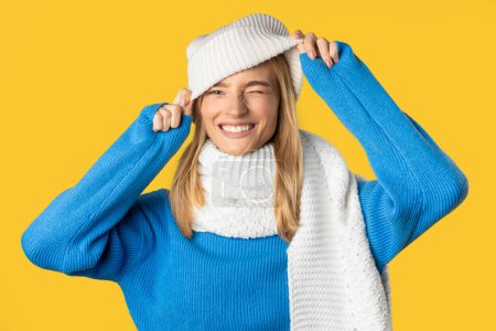 Eine Frau lächelt fröhlich, während sie ihren weißen Winterhut anpasst. Sie trägt einen blauen Pullover und einen dazu passenden weißen Schal und posiert vor einem leuchtend gelben Hintergrund.