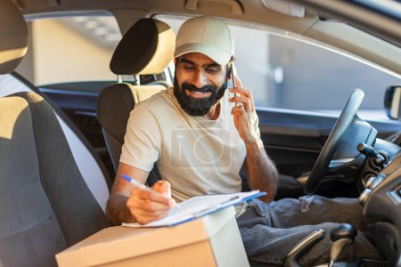 Indian delivery driver wearing a beige cap is seated inside his vehicle, talking on the phone and writing notes on a clipboard during the daytime.