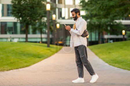 Indischer Mann läuft einen Bürgersteig entlang, während er in sein Handy vertieft ist, seine Aufmerksamkeit konzentriert sich auf den Bildschirm und nicht auf seine Umgebung, Multitasking, während er durch die städtische Umgebung navigiert.