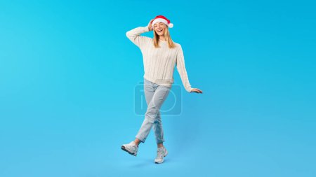 A young woman is standing on one leg, wearing a cozy sweater, jeans, and sneakers, all while donning a Santa hat. She has a joyful expression and her hand on her head