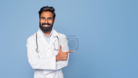 Ein fröhlicher indischer Arzt mit dunklem Bart und Stethoskop um den Hals zeigt lächelnd nach links. Er trägt einen weißen Mantel und steht vor einem festen blauen Hintergrund, Kopierraum