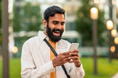 Indian bearded man is happily engaged with his smartphone while standing in a lush green park during a sunny afternoon. Lamps and modern buildings are visible in the blurry background
