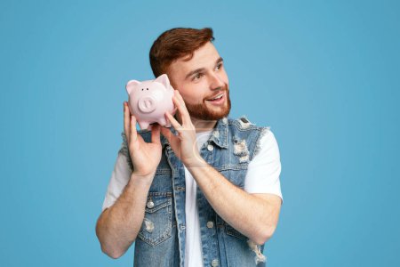 Dreamy millennial guy thinking about spending savings from moneybox, copy space, blue studio background
