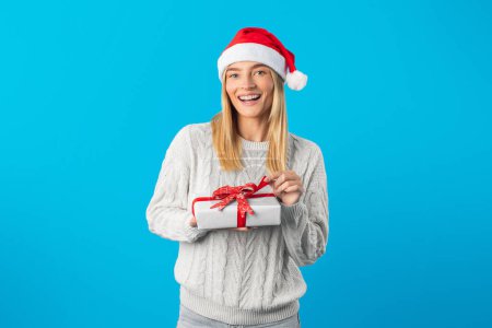 A happy woman stands against a vibrant blue backdrop wearing a Santa hat and a cozy sweater. She holds a neatly wrapped gift adorned with a red ribbon, showcasing holiday cheer.