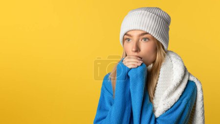 Une jeune femme portant un pull bleu et un bonnet blanc se tient pensivement sur un fond jaune audacieux. Son foulard blanc est drapé sur son épaule alors qu'elle semble perdue dans la pensée.