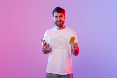 Photo for A man is holding a card in one hand and a cell phone in the other. He appears to be focusing on the phone screen while holding the card. The background is neutral and nondescript. - Royalty Free Image