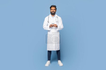 Ein indischer Arzt steht selbstbewusst in einem Studio, trägt einen weißen Mantel und lächelt herzlich. Er wirkt professionell und nahbar und suggeriert Bereitschaft, Patienten zu helfen.