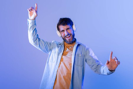 A young man enthusiastically dances to music, wearing headphones, in a vibrant, colorful room during nighttime. He looks happy and energetic.