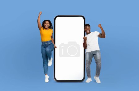 Two young black adults are joyfully celebrating while standing next to an oversized smartphone screen. The expressions on their faces show excitement and happiness, with their arms raised