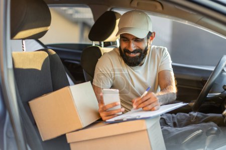 Indian bearded delivery driver wearing a cap is sitting inside his vehicle during the day. He uses his smartphone while holding a clipboard with parcels on the seat beside him