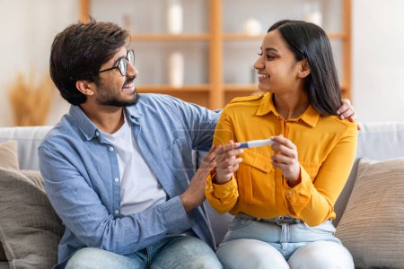 Photo for A joyful Indian couple is seated on a sofa in their cozy living room, smiling lovingly at each other while holding a positive pregnancy test. - Royalty Free Image