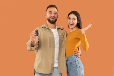 A cheerful couple is seen holding a set of car keys against a vibrant orange backdrop. The man displays the keys while the woman makes an enthusiastic gesture, indicating their joy and excitement