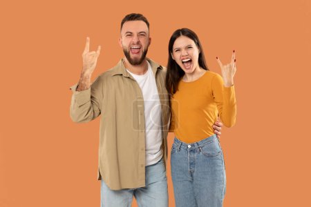 Ein fröhliches Paar macht begeistert rockige Handgesten und lächelt dabei breit. Der Hintergrund ist eine leuchtend orangefarbene Farbe, die ihren fröhlichen Ausdruck und ihre lässige Kleidung hervorhebt.
