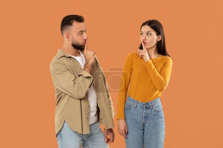 A young man and woman stand facing each other, both holding a finger to their lips, signaling for silence against a solid orange background. They seem to be engaged in a quiet conversation or secret.