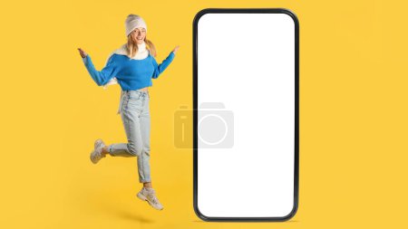 A cheerful young woman in a blue sweater and jeans joyfully jumps next to an oversized smartphone with a blank screen. The vibrant yellow background adds a lively touch to the scene.