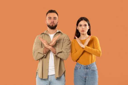 Un jeune homme et une jeune femme se tiennent côte à côte sur un fond orange, affichant un clair non geste en croisant les bras devant, semblent graves, indiquant une désapprobation ou un désaccord