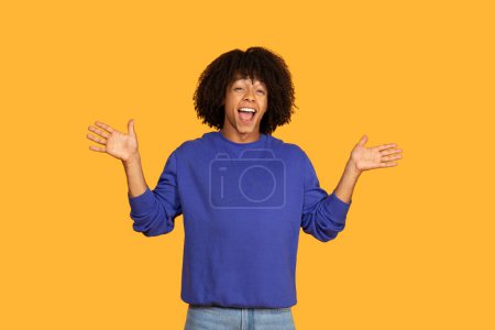 A young guy with curly hair is exuberantly expressing joy with an energetic pose. They are dressed in a blue sweater and jeans, standing in front of a bold yellow backdrop.