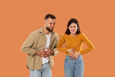 A young couple holds their stomachs, appearing to endure discomfort against an orange backdrop. Both are casually dressed, emphasizing their distress.