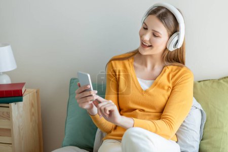 A teen girl is sitting comfortably on a couch, wearing white headphones and a yellow sweater, looking at her smartphone with a cheerful expression