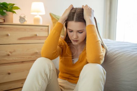 A teen girl, dressed in a yellow sweater and light pants, sits on her bedroom floor looking distressed with her hands on her head. Daylight illuminates the room from a nearby window.