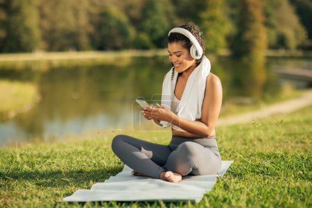 Athletic woman in sports attire is sitting cross-legged on a yoga mat, enjoying music with headphones as she relaxes by a lake in a serene park on a sunny day. She holds a smartphone