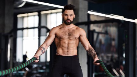 Muscular guy with naked torso exercising with battle ropes, crossfit training concept