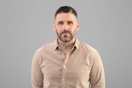 Ein Mann mit Kurzhaarschnitt und Bart steht vor einem schlichten grauen Hintergrund, trägt ein beiges Hemd und blickt direkt in die Kamera. Das Setting wirkt drinnen mit gleichmäßiger Beleuchtung.