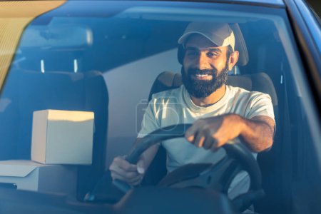 A cheerful Indian delivery driver navigates his vehicle through city streets early in the morning. He is seated behind the wheel, with packages visible beside him