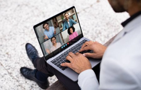 A businessman is participating in a video conference call on his laptop while sitting outdoors in an urban environment. Five colleagues appear on the screen in a professional, virtual meeting.